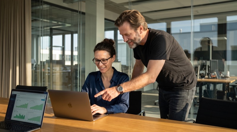 Katja Müller und Stefan Richter prüfen das Navigator-Dashboard in einem gläsernen Konferenzraum, während warmes Morgenlicht hereinfällt.