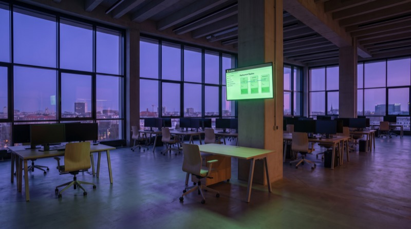Die Entwicklungsfläche von Pixel Spree in einer tiefvioletten Berliner Sommerdämmerung. Das Büro ist leer, mit aufgeräumten Schreibtischen, dunklen Monitoren und einem einzigen Wandschirm, auf dem ein durchgehend grün leuchtender Jenkins-Pipeline-Block zu sehen ist.